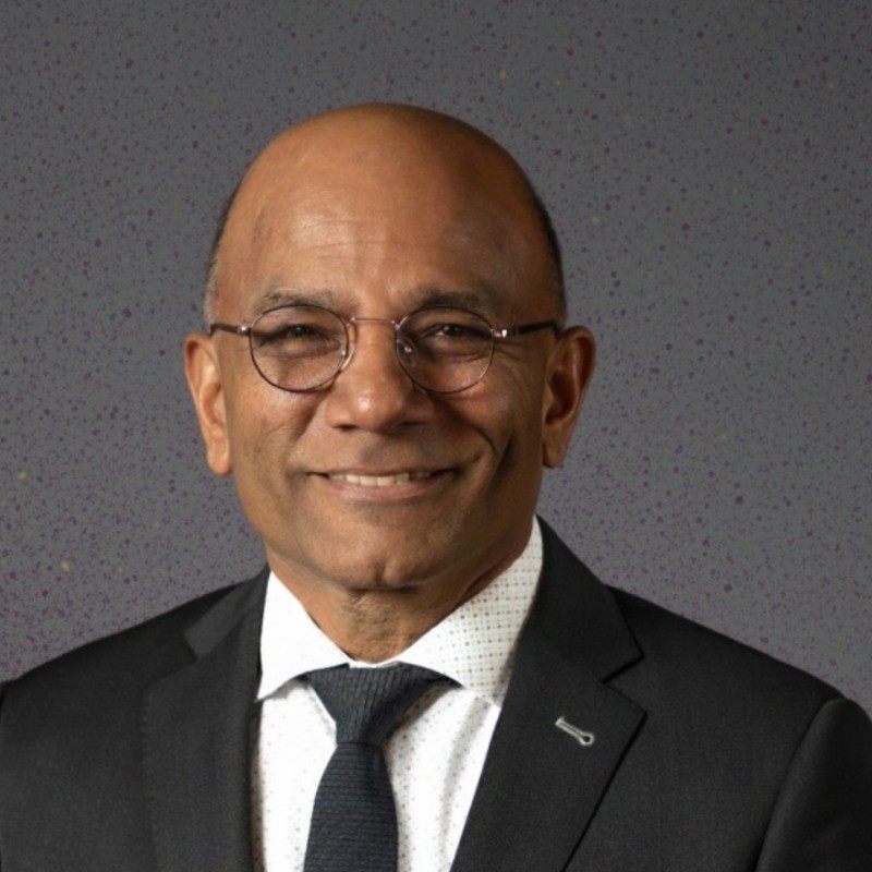 Smiling man in a dark suit, white shirt, and navy tie, wearing round glasses; professional headshot against a speckled gray background.