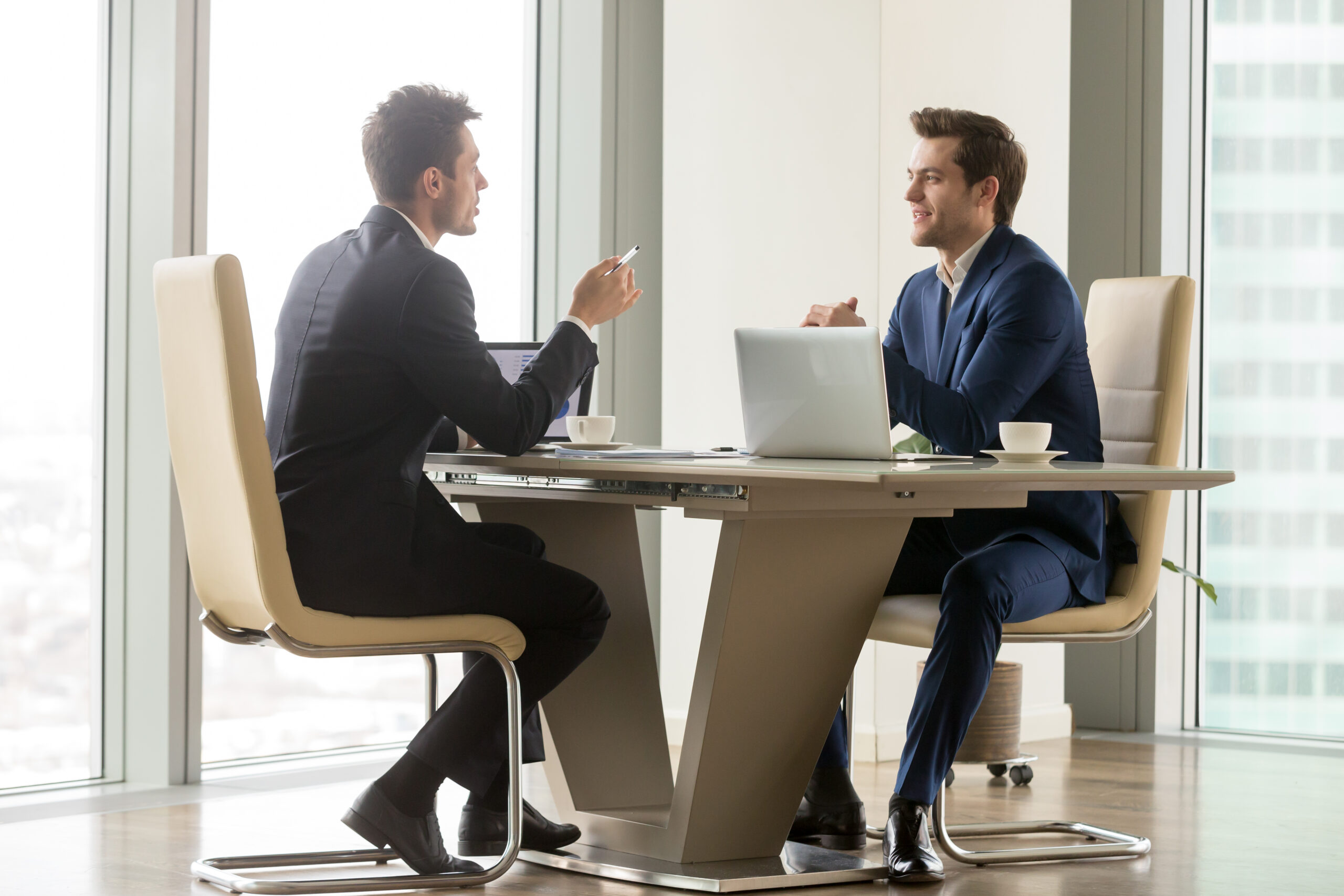 Two handsome businessmen sitting in comfortable chairs at desk with laptops in meeting room. CEO making important negotiation about companies partnership or corporate merger. Financiers planning deal