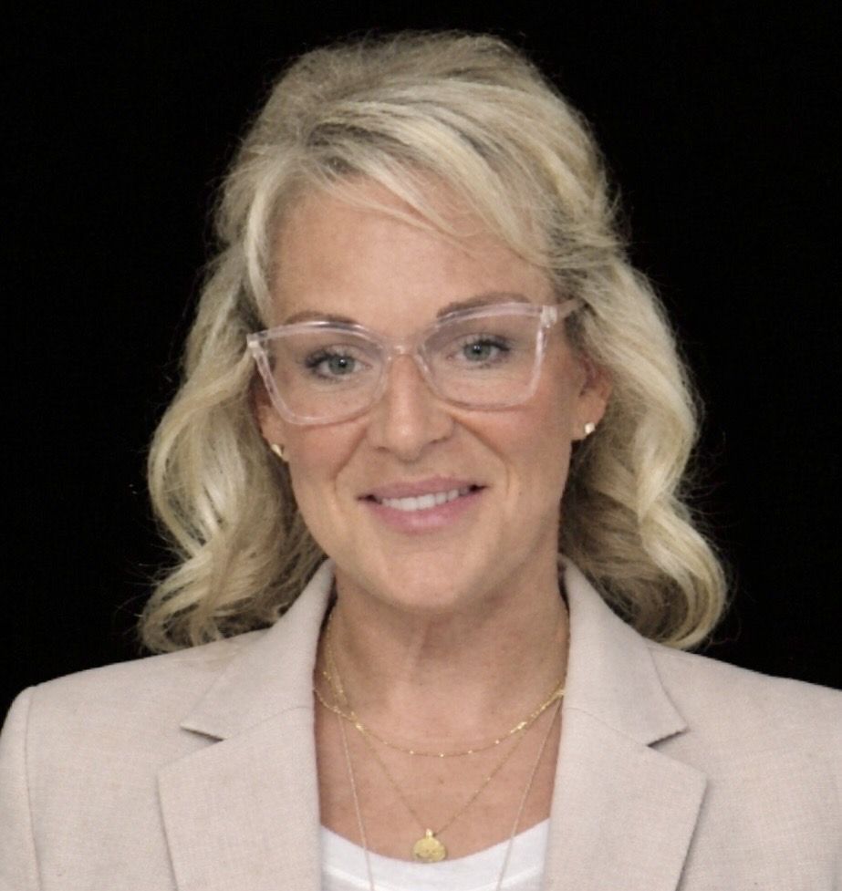 Portrait of a smiling woman with blonde hair and clear eyeglasses, wearing a beige blazer against a black background.